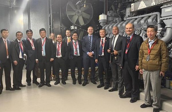 A group of thirteen people in formal attire in front of large data centre equipment including a fan as part of the cooling system. 