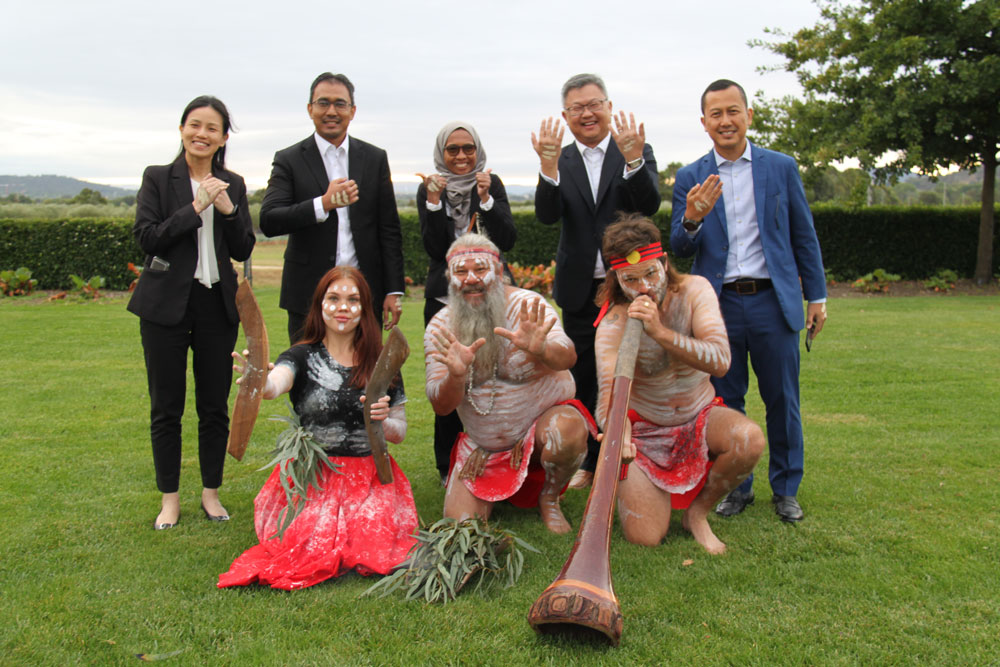 Two women and three men stand on a green lawn, dressed in business suits. In front of them, three Indigenous performers pose with traditional instruments, wearing white ceremonial body paint.