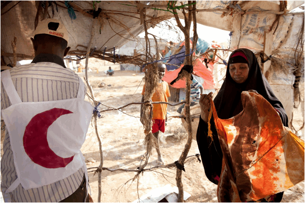 A male Red Cresent worker is helping an African woman wearing a black abaya to construct a makeshift tent from sticks and fabric.
