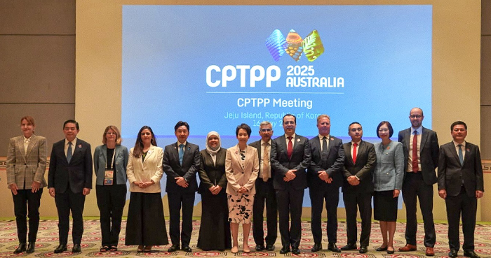 14 people standing side by side in a large room, with a screen displaying text 'CPTPP 2025 Australia; CPTPP Meeting, Jeju Island, Republic of Korea' in the background