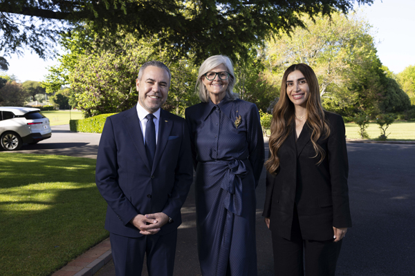 Three people standing together outdoors in a garden, with trees and lawns in the background.