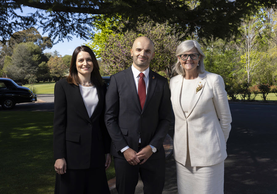 Three formally dressed adults posing outdoors on a tree-lined property. Center man wears black suit and red tie, flanked by woman in black suit and woman in white suit with glasses and brooch, suggesting an official or ceremonial gathering.