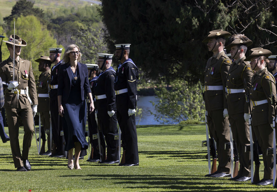 A formal military ceremony on a grassy lawn, with uniformed personnel standing in formations beside a woman in a navy suit. Soldiers wear khaki or dark navy uniforms, brimmed hats, medals, and ceremonial belts, with trees and a lake creating a scenic outdoor setting.