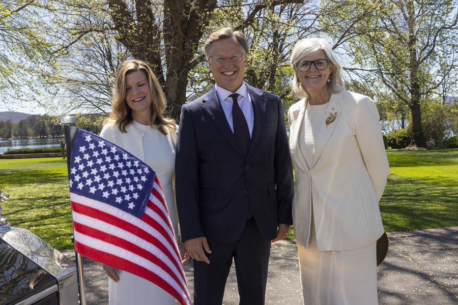 Three formally dressed adults posing outdoors beside a U.S. flag, with a lake, trees, and lawn behind them. Center man wears a dark suit and tie, flanked by two women in light outfits, suggesting a ceremonial or official gathering.