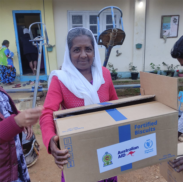 An individual outdoors holding a cardboard box labeled “Fortified Biscuits” with logos for “Australian Aid,” the WFP, and a local emblem. The setting includes a building with windows and gardening tools in the background, suggesting a distribution point for aid supplies.