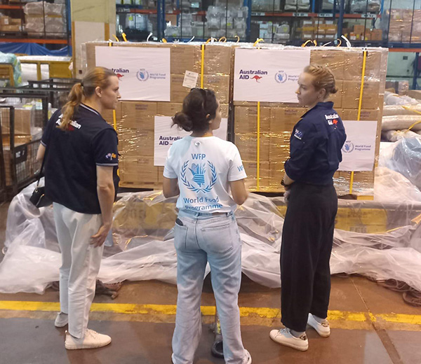 Three individuals standing in a warehouse, observing large pallets wrapped in plastic and secured with yellow straps. The pallets have labels displaying “Australian Aid” with a kangaroo logo and the World Food Programme (WFP) emblem. The background shows shelves stacked with boxes and supplies.