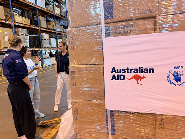 A close-up view of stacked boxes wrapped in plastic inside a warehouse. A large label on the boxes reads “Australian Aid” with a kangaroo logo and the WFP emblem. Three individuals are standing nearby, engaged in conversation. Additional boxes and shelving units are visible in the background.