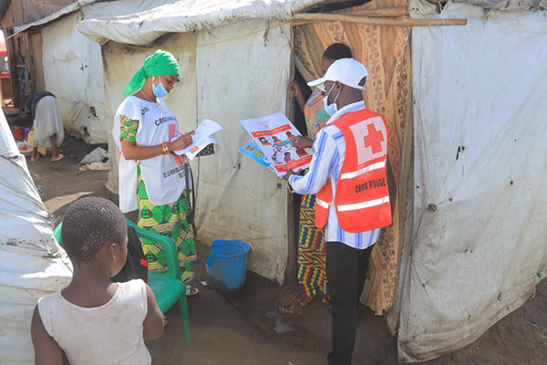 Red Cross volunteers distribute Ebola awareness materials and collect information from residents outside temporary shelters in a camp setting, while a child stands nearby.