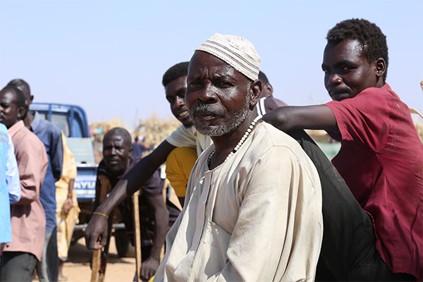 A group of people gathered outdoors in a dry, open area with clear blue sky in Sudan. Some individuals are sitting while others stand nearby. A vehicle is visible in the background along with humanitarian supplies. 