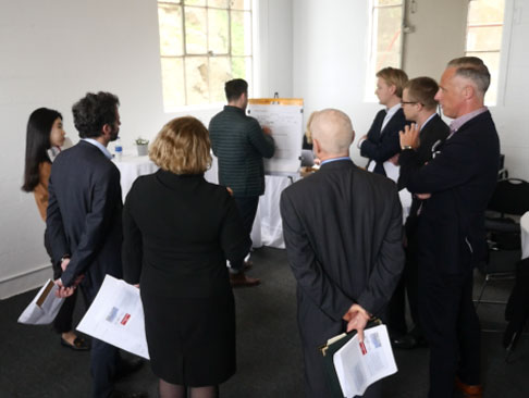Photo of a group of people standing around watching a person write on a piece of paper.