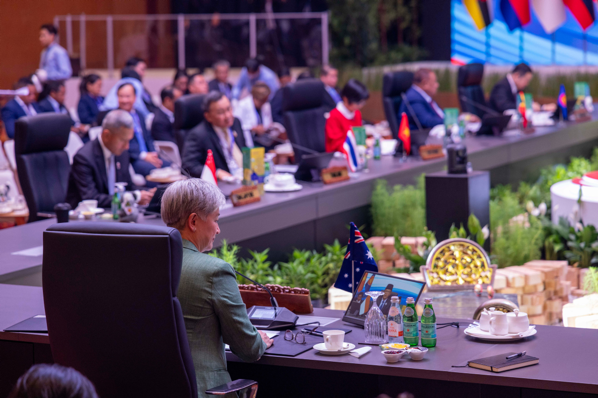 Photograph of an international summit with suited delegates seated around long conference tables, reviewing documents and laptops. National flags, microphones, nameplates, bottled water, ceremonial table settings indicate formal diplomatic event.