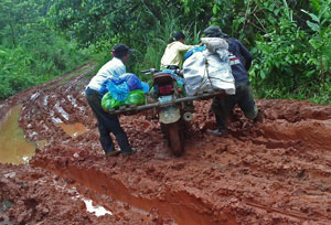 Three people trying to move a motorcycle carrying large bags along a very muddy road criss-crossed by deep wheel ruts