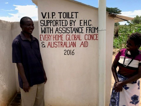 Man and woman standing in front of toilet. The toilet wall has wording: VIP toilet supported by EHC with assistance from Every Home Global Concern and Australian Aid 2016.