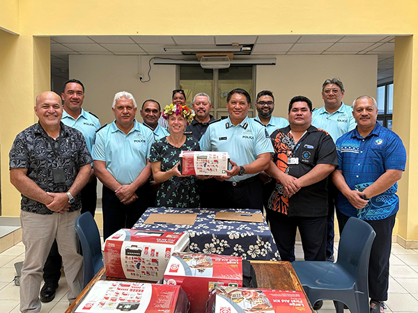 Group of twelve people indoors, some wearing light blue 'POLICE' shirts, standing behind a table with floral cloth and covered in first aid kits, one person holding a first aid kit, suggesting a law enforcement or community service event.