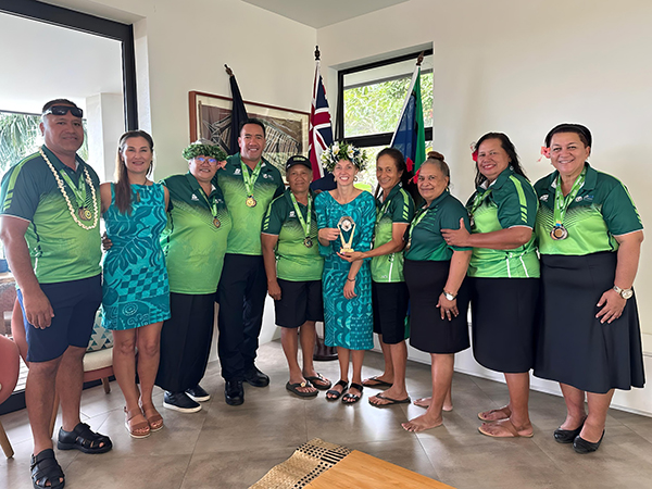 Group of ten people indoors wearing green shirts and black bottoms, some with medals, one holding a trophy, standing in front of two flags including the Union Jack, at a celebratory or award event