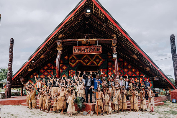 Group of people in traditional and formal attire posing in front of a culturally significant building with a triangular roof and intricate designs, labeled 'MAUATUMUARI', during a community celebration in Vanuatu.