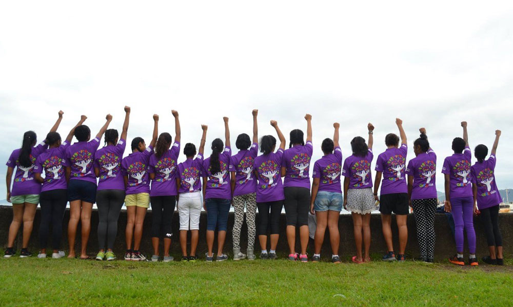 Group of young women facing away from the photographer all pumping a fist into the air and wearing their purple matching shirts. 