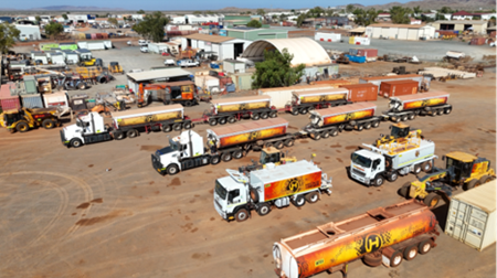 Aerial view showing an industrial transport yard with multiple fuel tankers, heavy machinery, shipping containers, and warehouses. Orange-and-white tanker trucks dominate foreground and center, while loaders, trailers, parked vehicles, and low-rise buildings extend across background.