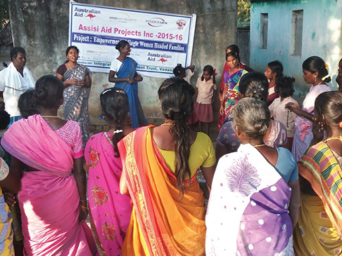 Group of women standing in front of Assisi Aid Projects Inc 2015-16 sign, branded with the Australian Aid logo