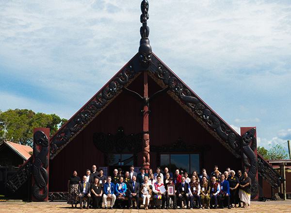 A large group of people stand and sit in rows in front of an ornately carved timber meeting house with a steep triangular gable, decorative carved figures along the roofline, and a central carved post. The building is dark red-brown wood, set outdoors under a blue sky.