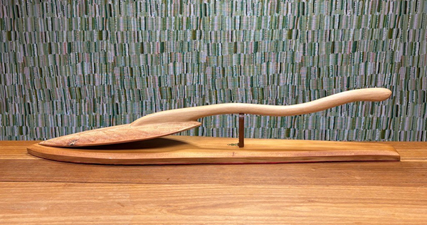 A long, smooth, carved wooden object with a curved handle and tapered blade-like form rests horizontally on a wooden base atop a table, displayed against a patterned green woven backdrop.