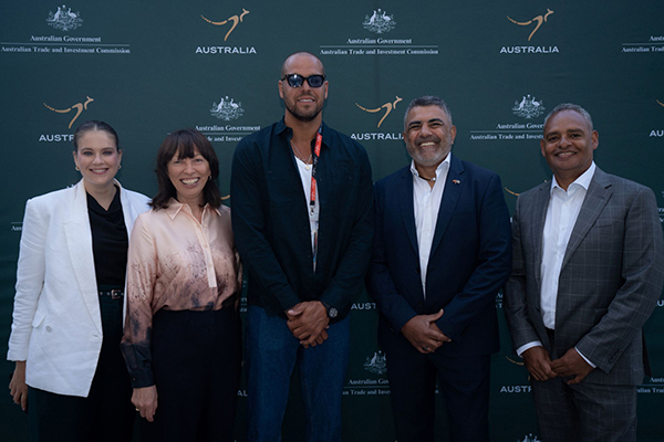 A group of five people standing in front of a green backdrop branded with the Australian Government and Austrade logos. They are dressed in business or smart-casual attire, standing side by side and facing the camera.