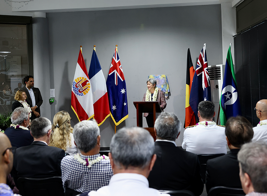 Foreign Minister Senator Wong at a podium, three flags on either side of her, presenting to a group of people seated in chairs.