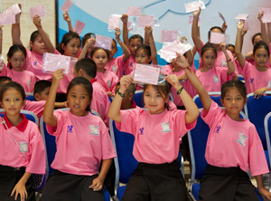Large group of smiling girls and boys in pink t-shirts, waving their pink vaccination certificates in the air