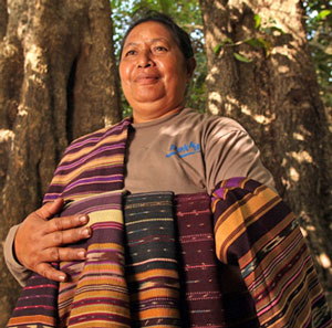 Indonesian woman standing outside in front of two large trees displaying her woven garments.