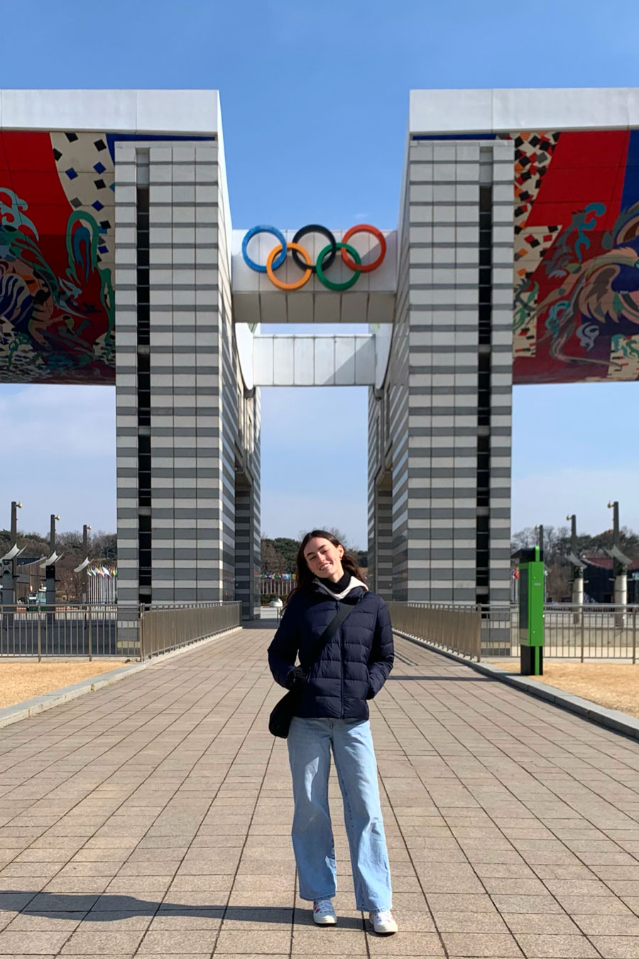Person standing in front of large sculptural entrance and Olympic rings logo 