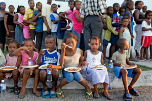 Photo of children eating a meal on their laps, with a queue of people in the background