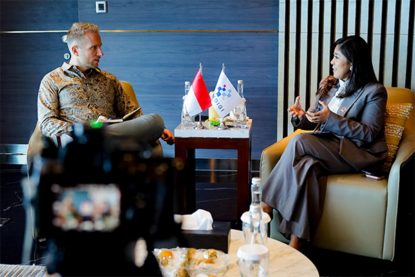 Two people seated in armchairs during an interview, speaking across a small table with two flags, water bottles, and notes; a camera and blurred foreground objects frame the scene.