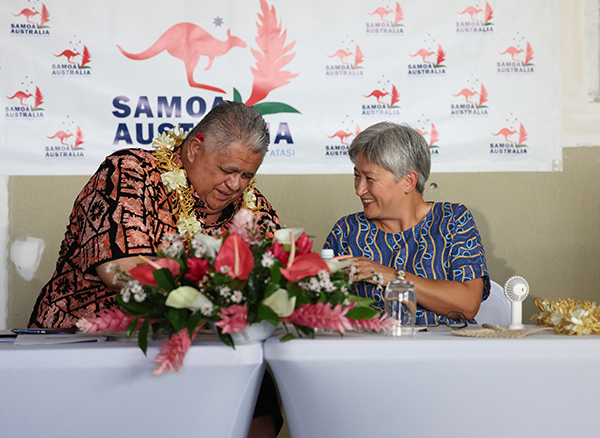 Two people seated at a table sign documents in front of a “Samoa Australia” backdrop, with a large floral arrangement in the foreground, water glasses, and leis worn during a formal event setting.