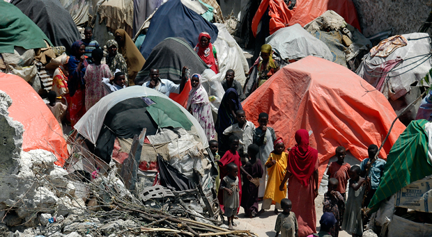A number of African people, mostly women and children, are walking through an area of rubble crowded with makeshift tents