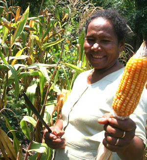 A woman smiles holding a cob of corn in a field.