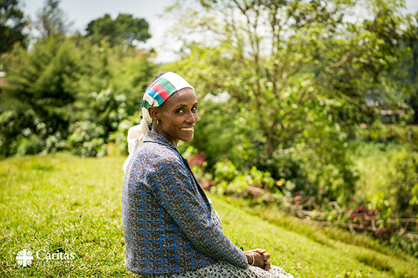 A person is sitting on a grassy hillside in an outdoor setting, wearing a patterned blue jacket and a headscarf with green, white, and red stripes. The background features lush greenery, trees, and a slightly blurred landscape, suggesting a rural or natural environment. The Caritas Australia logo is visible in the bottom left corner of the image.