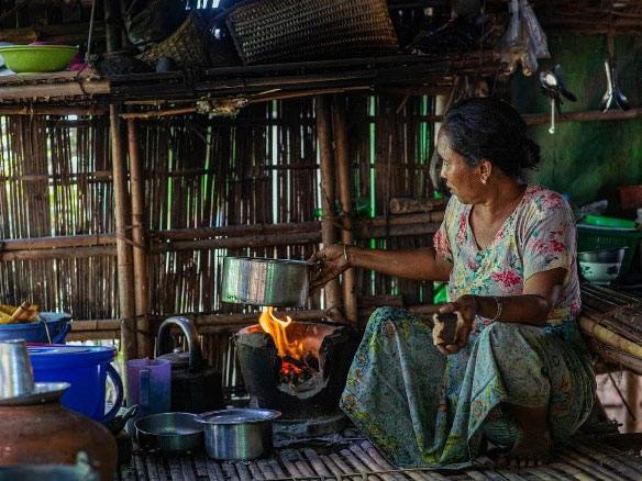 Woman in Myanmar using fuel sticks to cook food on stove.