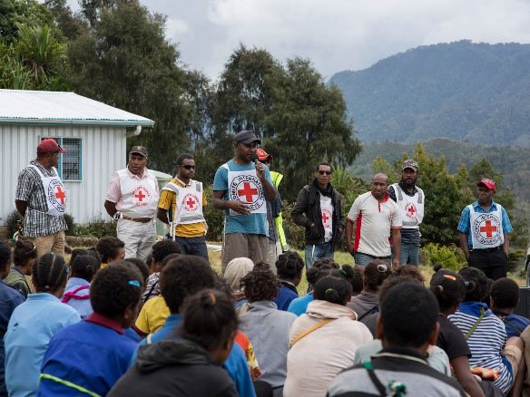 The Papua New Guinea Red Cross Society which is a group of men is discussing their work in front of a crowd of elementary school
