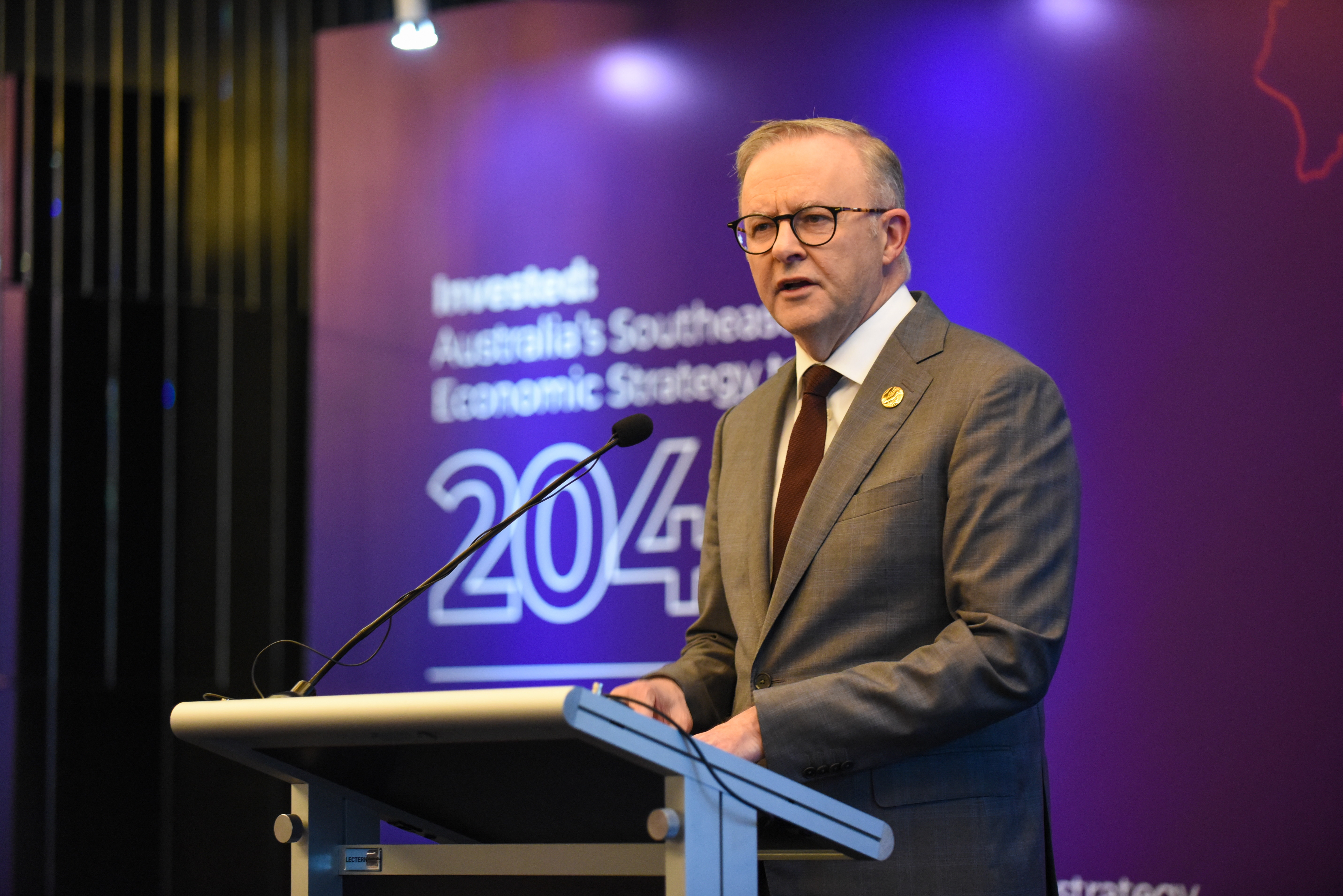 Photograph of a suited man speaking at a lectern with a microphone. Purple backdrop displays text about Australia’s Southeast economic strategy and a large “2040” graphic.