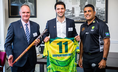 Photo of three men, one is holding a cricket stump covered in indigenous art and another is holding a jersey that says Bripumyarrimin and the number eleven.
