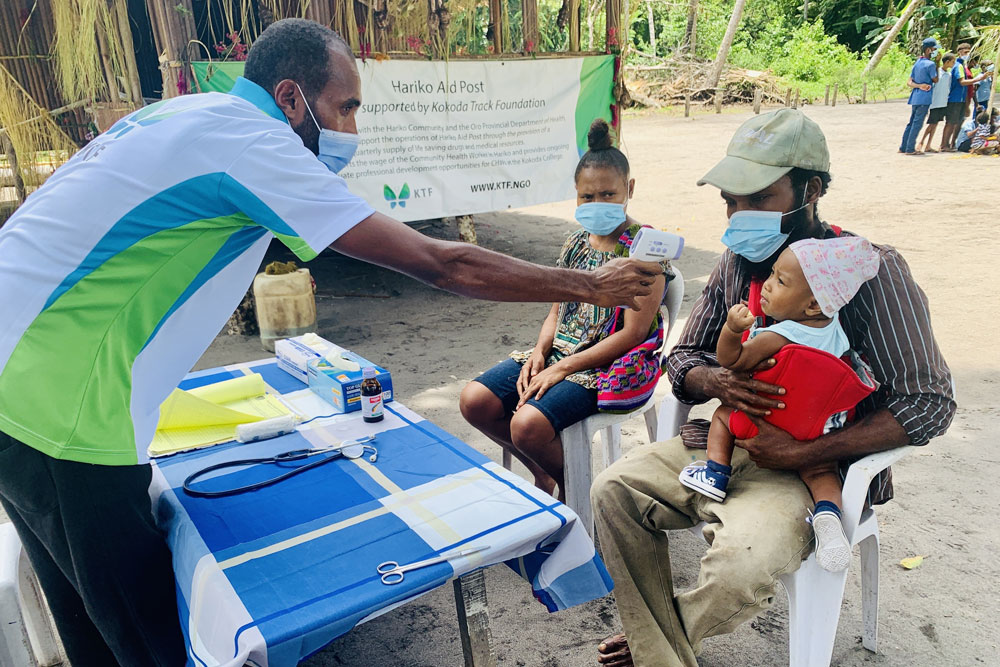 Community Health Worker doing a temperature check on a baby sitting in the fathers lap. 