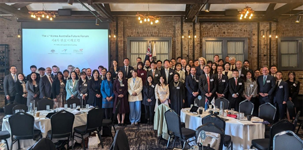 Group photo of formally dressed participants at the 3rd Korea-Australia Future Forum, taken in a conference room with brick walls, round tables, and a projection screen displaying the event title in English and Korean