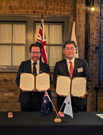 wo formally dressed individuals holding open documents in front of Australian and South Korean flags, standing at a table with miniature flags—likely during an official signing ceremony.