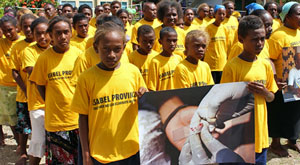 Large group of children in matching yellow t-shirts, the the front row holding a poster showing a finger-prick blood test for malaria