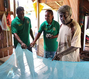 Two men and a woman inspect a blue mosquito net in an open residence with no screens or windows.