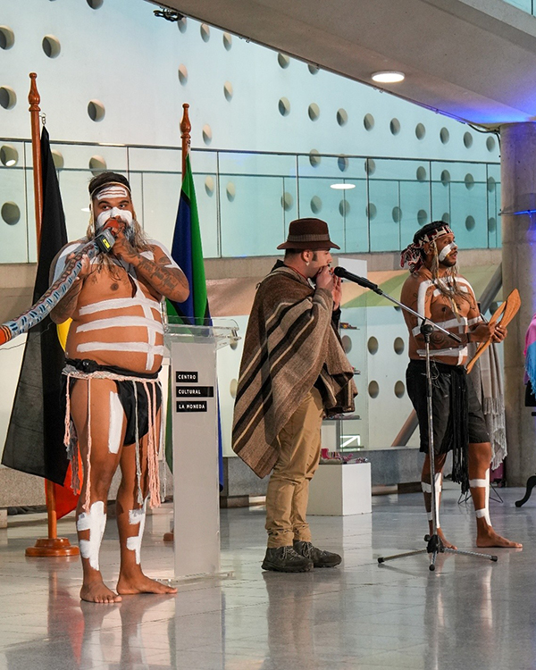 An indigenous Australian playing a didgeridoo while painted in traditional body paint, beside two indigenous Chilean musicians.