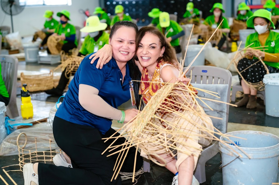 Photograph of two smiling women participating in a traditional bamboo weaving workshop, surrounded by workers in bright green uniforms. Bamboo strips, woven baskets, plastic buckets, and craft tools fill workshop, highlighting hands-on cultural craftsmanship and collaborative activity.