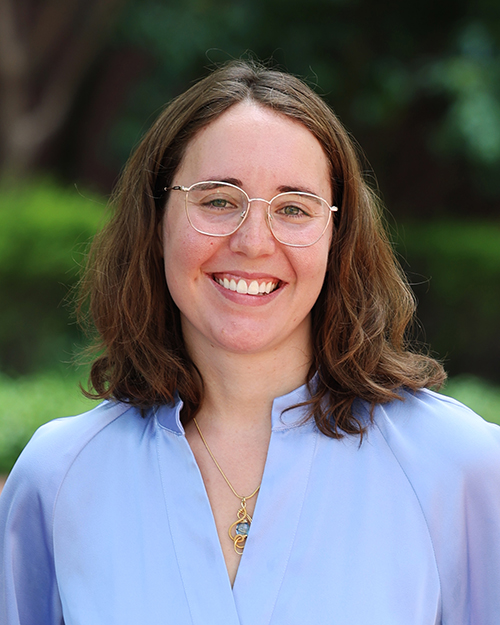 A person dressed in a light blue blouse with a pendant necklace, standing in an outdoor garden setting.