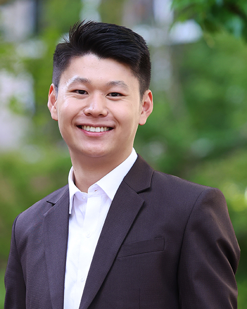 A person wearing a dark suit jacket over a white collared shirt, standing outdoors with green foliage in the background.
