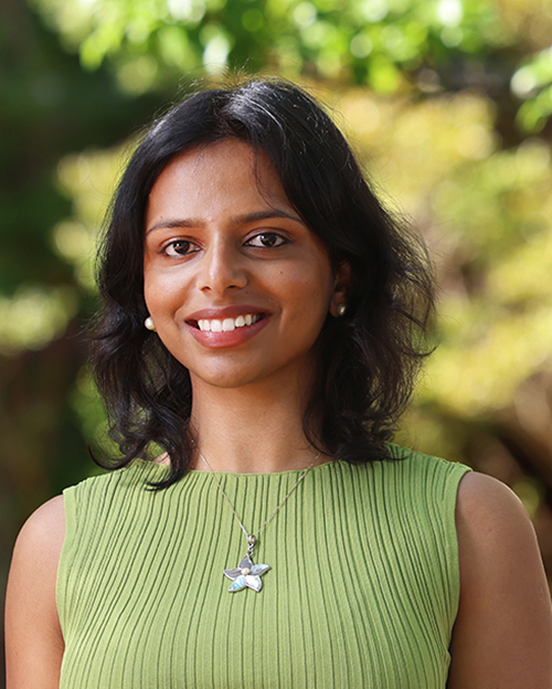 A person in a sleeveless, ribbed light‑green top and a star-shaped necklace, standing in a sunlit outdoor environment with trees in the background.